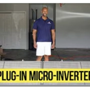 Man in a navy polo and white shorts stands in a garage behind a table with a bold 'Plug-in Micro-Inverter' sign.