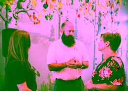 Teddy-Ronald McDonald - APsystems USA | The global leader in multi-platform MLPE technology - APsystems USA | The global leader in multi-platform MLPE technology Bearded man in a white polo speaks with two women in front of a colorful tree mural indoors, mid-conversation.