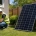 Worker in hard hat and high‑visibility vest crouches by wall outlet, wiring a cable during solar panel installation in a yard.