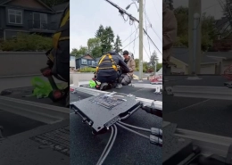 Two technicians in safety harnesses kneel on a flat residential roof, working with electrical equipment and cables near a utility pole in the background.