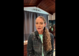 Woman with long curly hair wearing a denim jacket speaks to the camera in a cozy room with a wooden ceiling, gray curtains, and a vase on a table in the background.