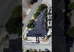 Aerial view of a multi-building complex with dark gray roofs, surrounded by trees, at a street corner with parked cars nearby.